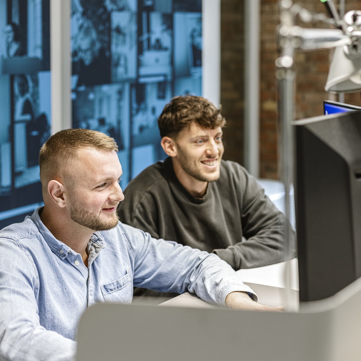 Zwei männliche Mitarbeiter von HOCHZWEI blicken gemeinsam auf einen PC und lächeln