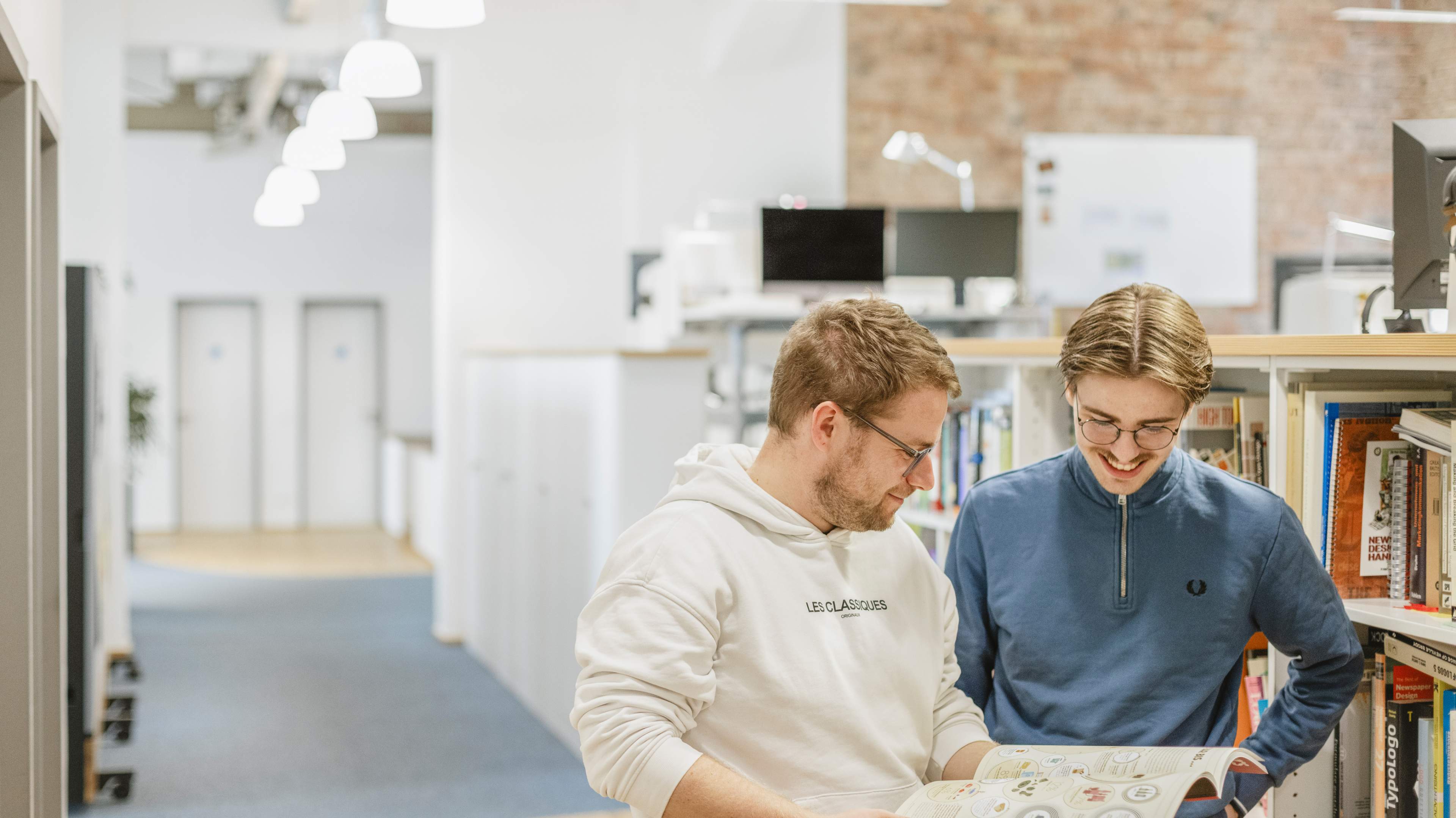 Zwei männliche Mitarbeiter von HOCHZWEI stehen im Flur und blättern durch einen Printkatalog