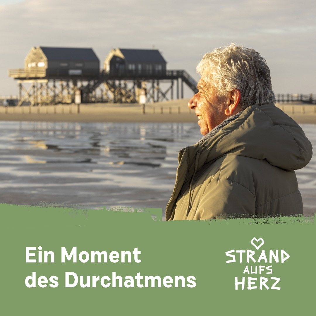 Ein älterer Mann steht am Strand vor Pfahlbauten unten im Bild der Schriftzug ein Moment des Durchatmens Strand aufs Herz