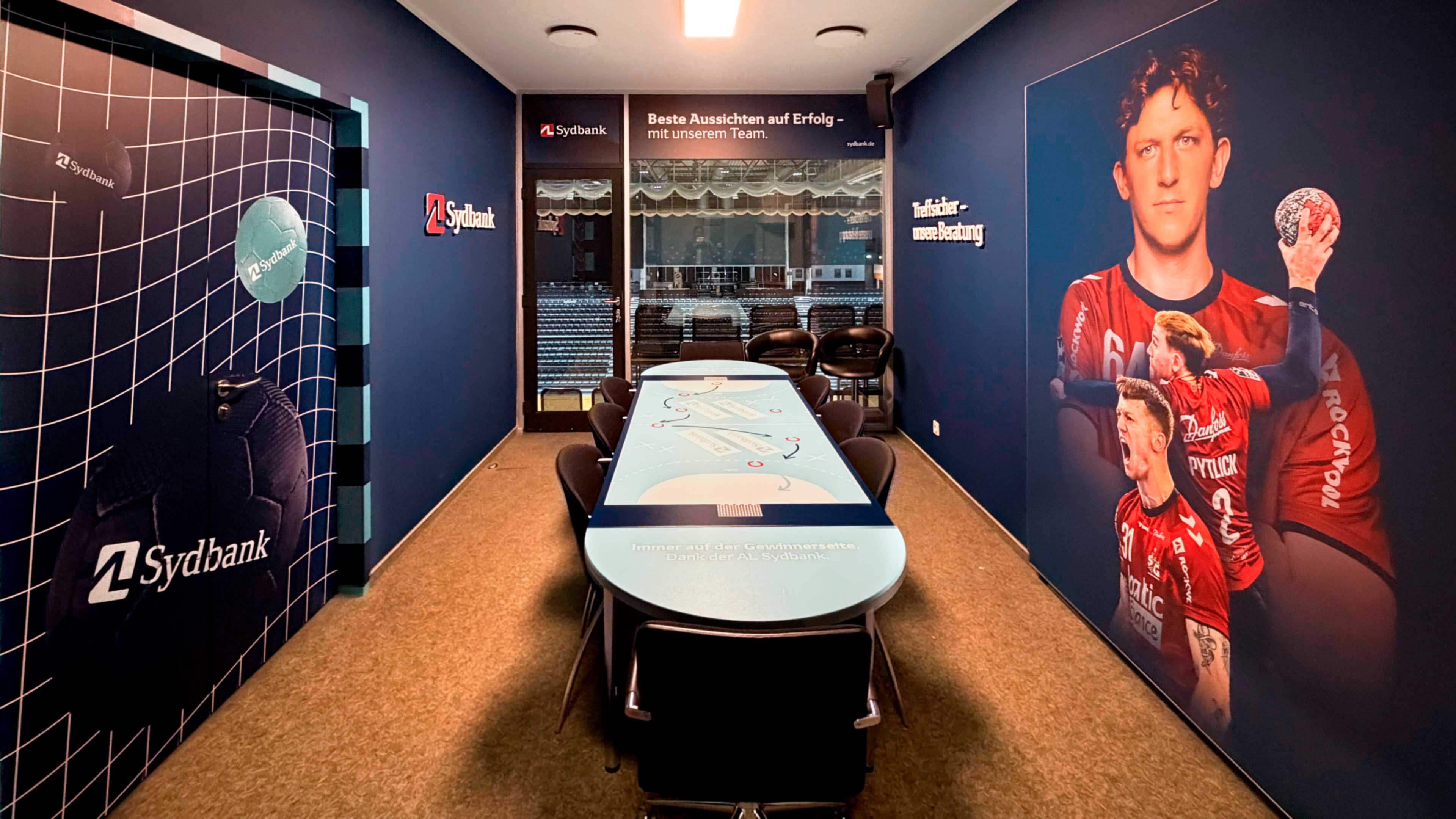 Eine Loge in der GP Joule Arena, mit dunklen Wänden, darauf ein Handballspieler in rotem Hemd