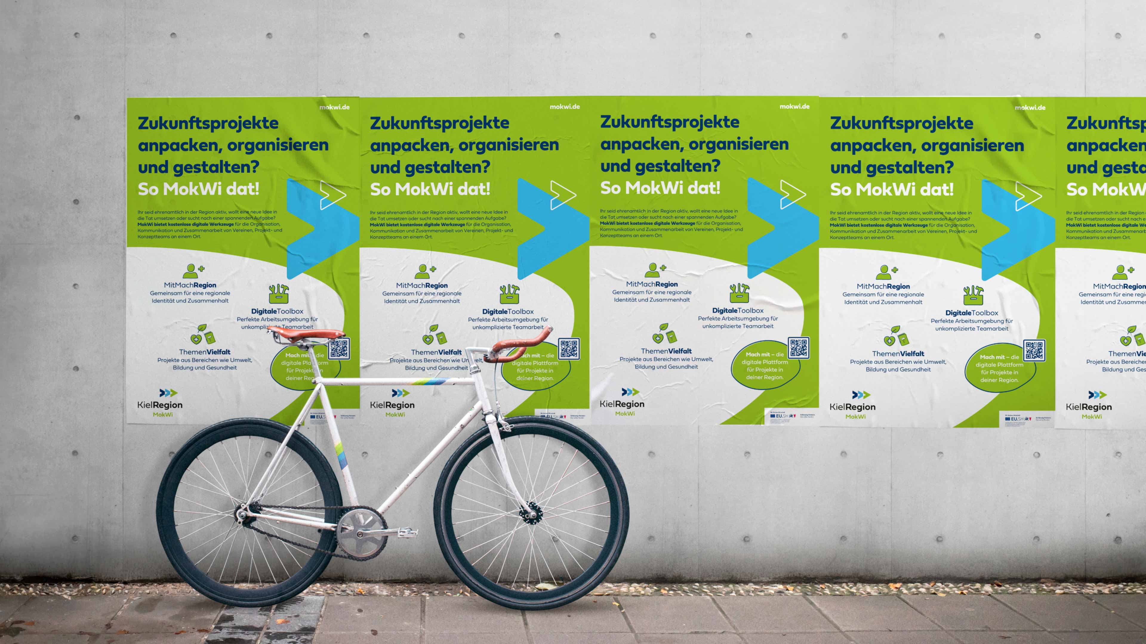 Eine graue Außenwand mit fünf MokWi Plakaten in einer Reihe. Darauf steht Zukunftsprojekte anpacken und organisieren und gestalten? So MokWi dat. Darunter in klein viele weitere Informationen. Vor der Wand steht ein Fahrrad. Die Plakate sind weiß-hellgrün mit einem hellblauen Pfeil nach rechts.