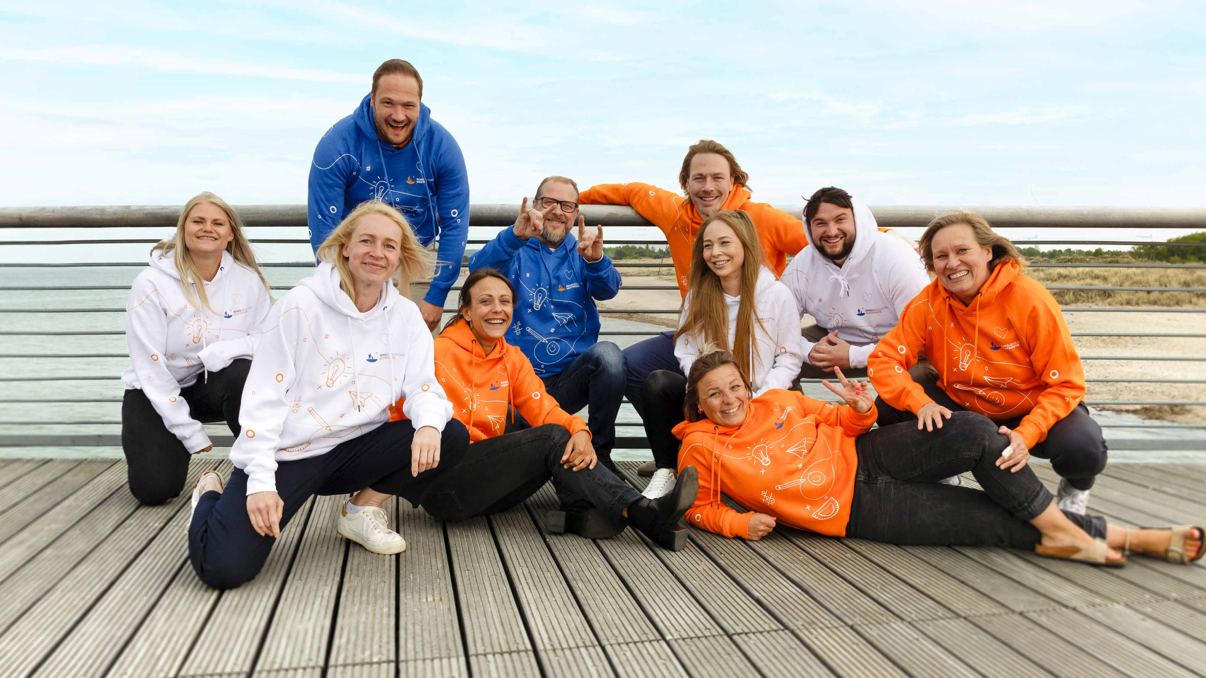 Das Team der Schulbegleitung Westküste in den neuen Hoodies in jeweils Weiß, Orange und Blau bei einem Fotoshooting auf einem Steg am Strand.