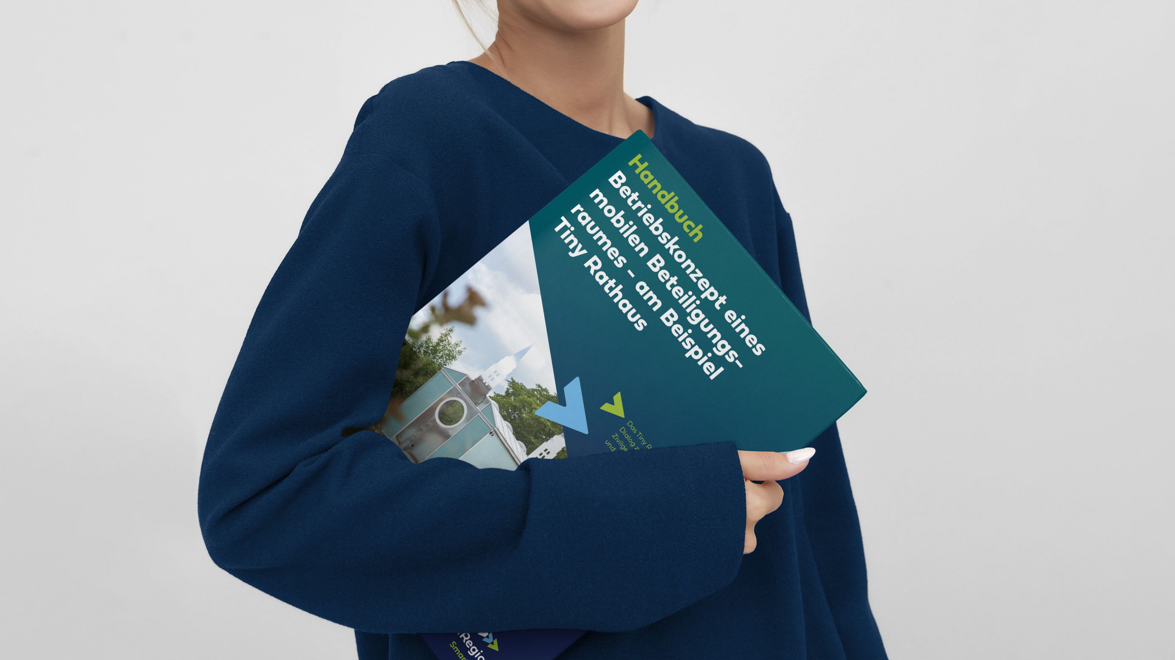 Eine Person hält ein Handbuch mit dem Titel Betriebskonzept eines mobilen Beteiligungsraumes - am Beispiel Tiny Rathaus