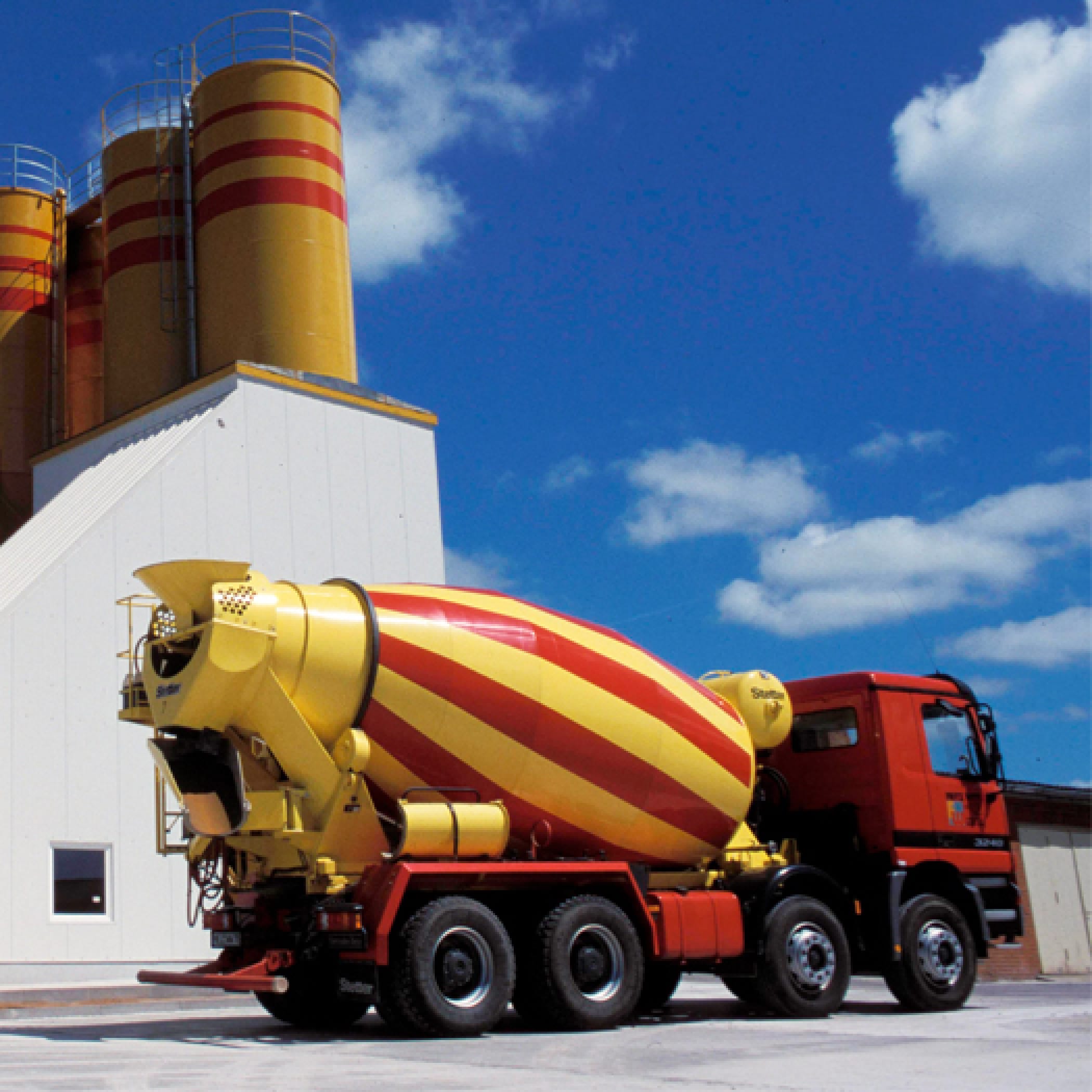 Ein gelb-roter Betonmischer vor einem hohen Silo-Gebäude