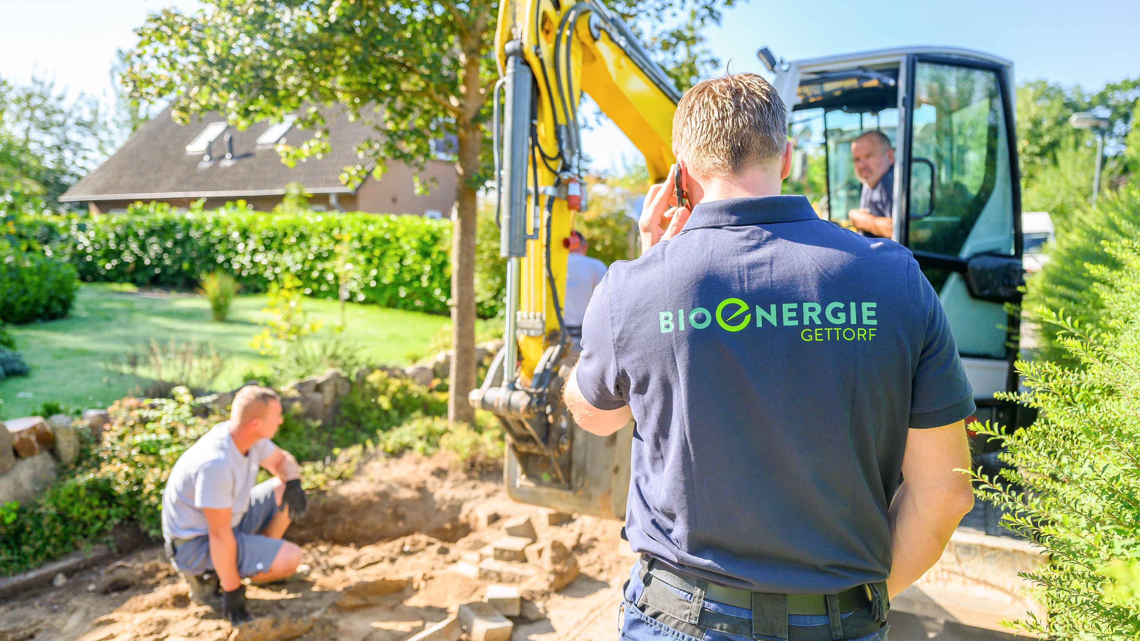 hochzwei_bioenergie_slider_02.jpg Rechteckiges Bild einer Person mit blauem Poloshirt mit Bioenergie Gettorf Logo steht vor einem kleinen Bagger und telefoniert