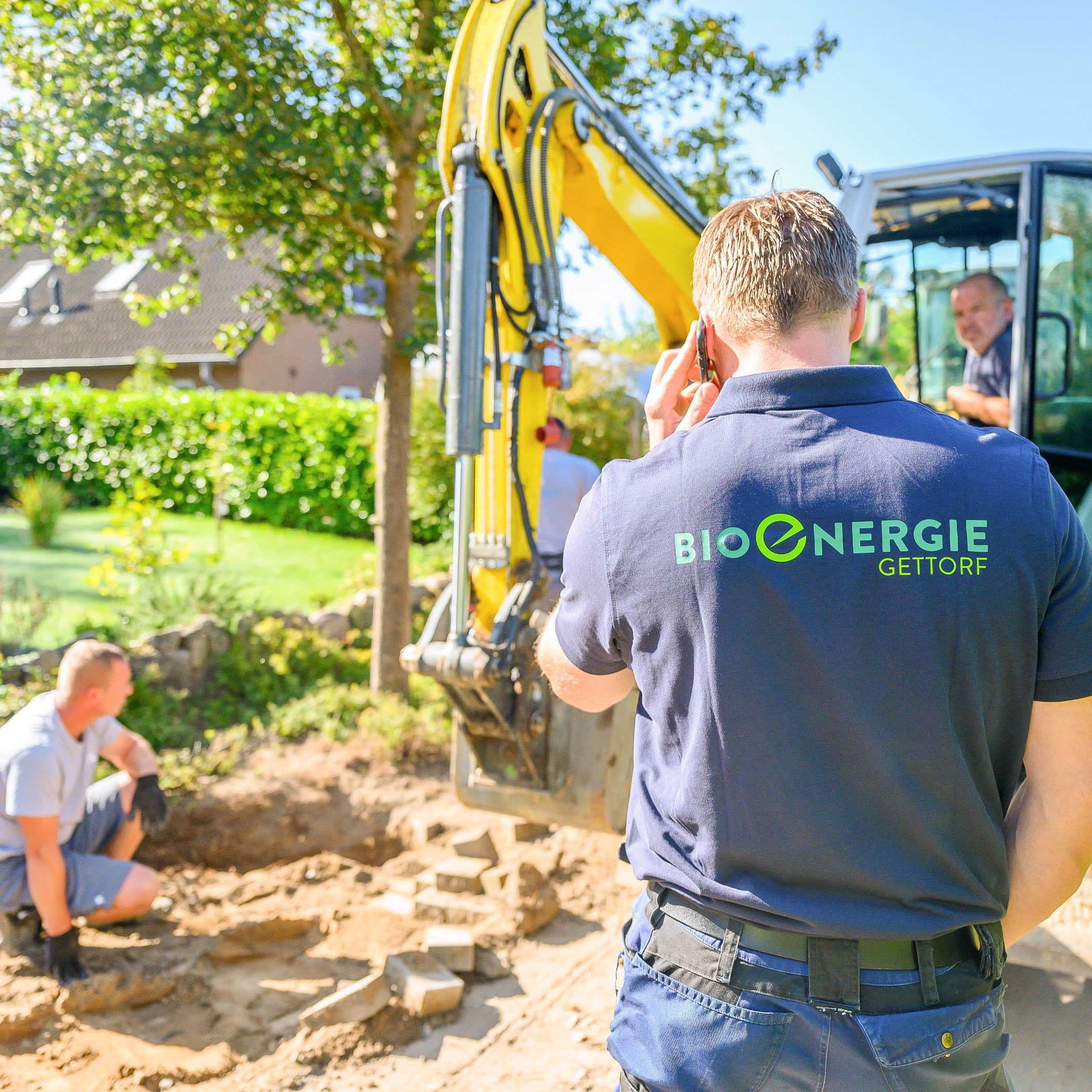hochzwei_bioenergie_teaser_07.jpg Quadratisches Bild einer Person mit blauem Poloshirt mit Bioenergie Gettorf Logo steht vor einem kleinen Bagger und telefoniert