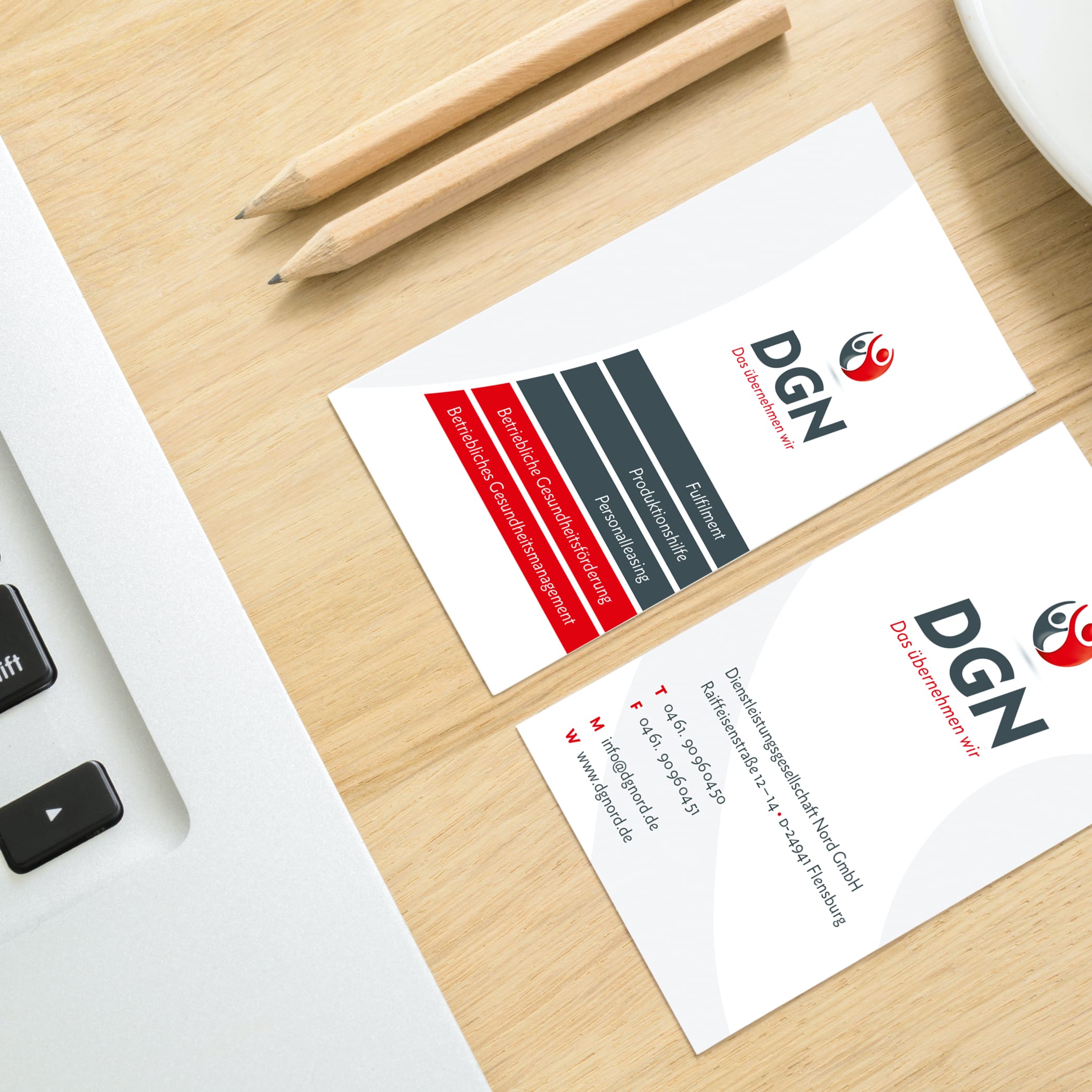 Zwei Visitenkarten für DGN liegen auf einem Holztisch, zwei Bleistifte daneben