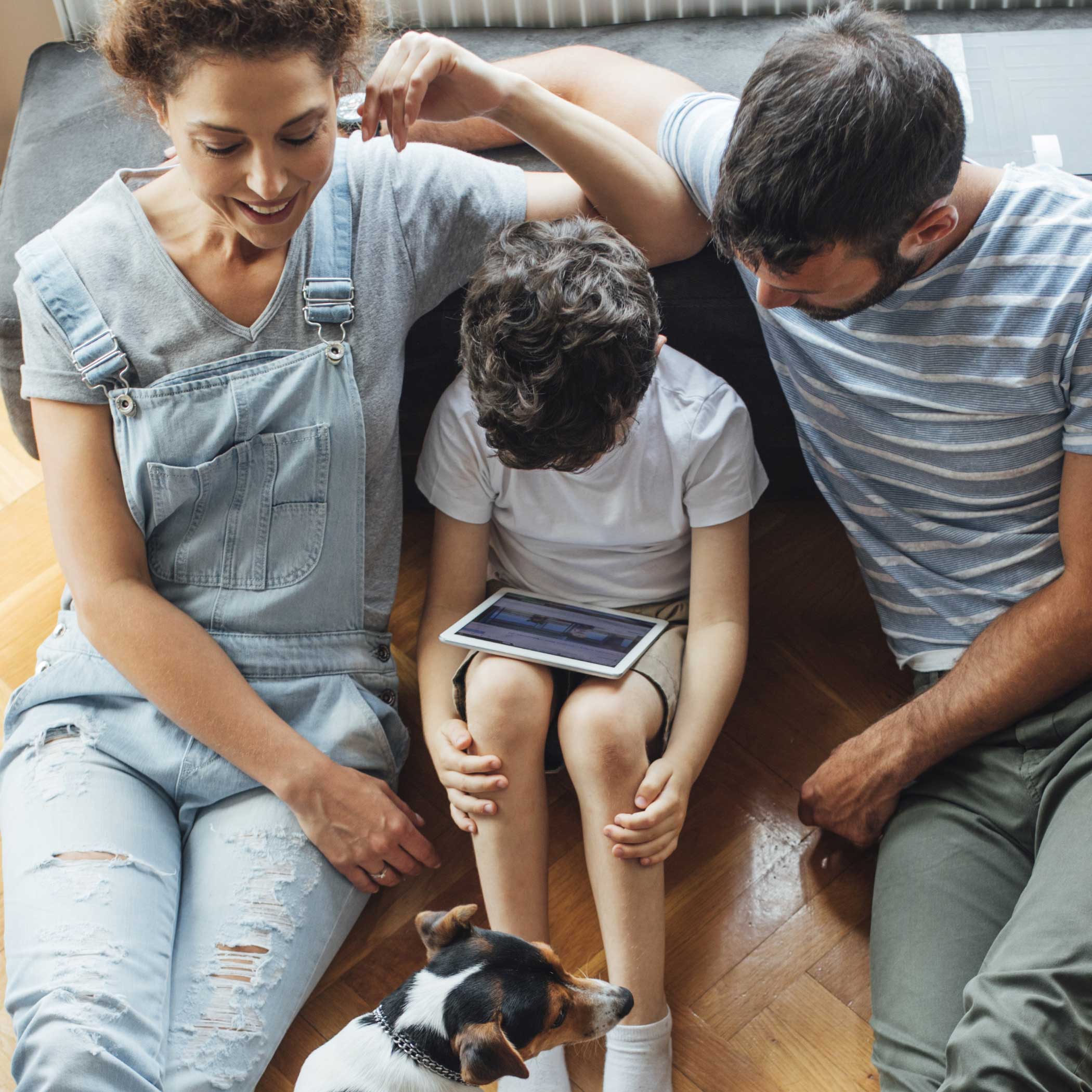 hochzwei_domcura_teaser_04.jpg Aufnahme von schrägoben: Mutter, Kind und Vater sitzen auf dem Fußboden und schauen auf Tablet auf dem Schoß des Kindes