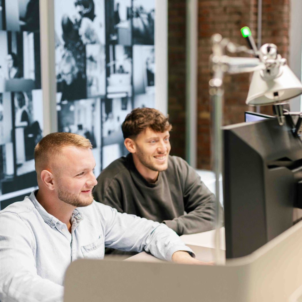 Zwei männliche HOCHZWEI-Mitarbeiter blicken gemeinsam und lächelnd auf Bildschirme