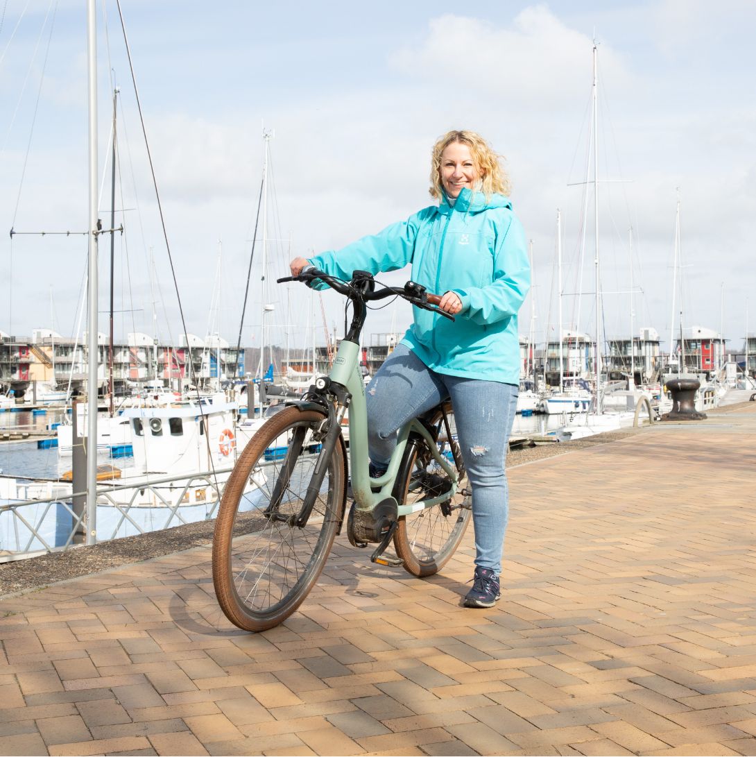 h2_Arbeitgeber_teaser_04.jpg Eine Hochzwei Angestellte mit einem Fahrrad an der Flensburger Fördepromenade. Sie lächelt in die Kamera.