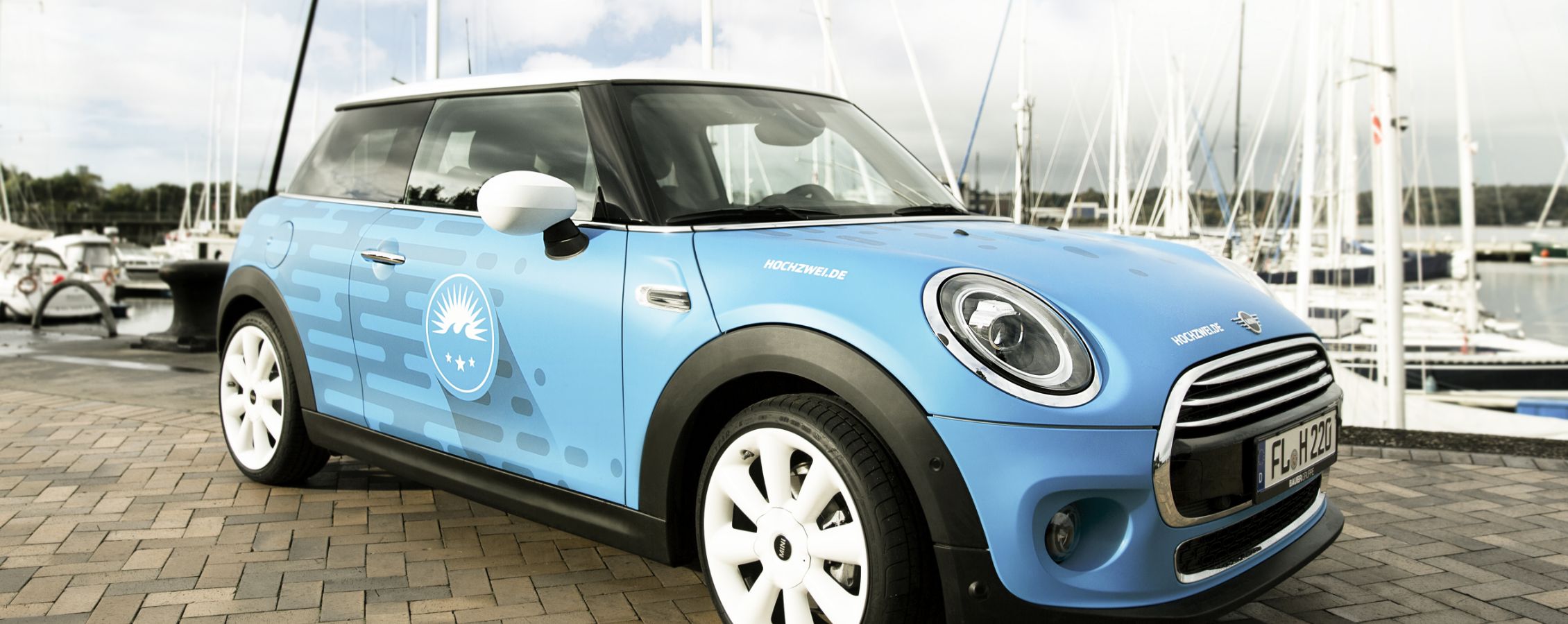 h2_Arbeitgeber_teaser_06.jpg Ein Auto der Marke Mini in hellblau mit Hochzwei-Druck an der Flensburger Fördepromenade.