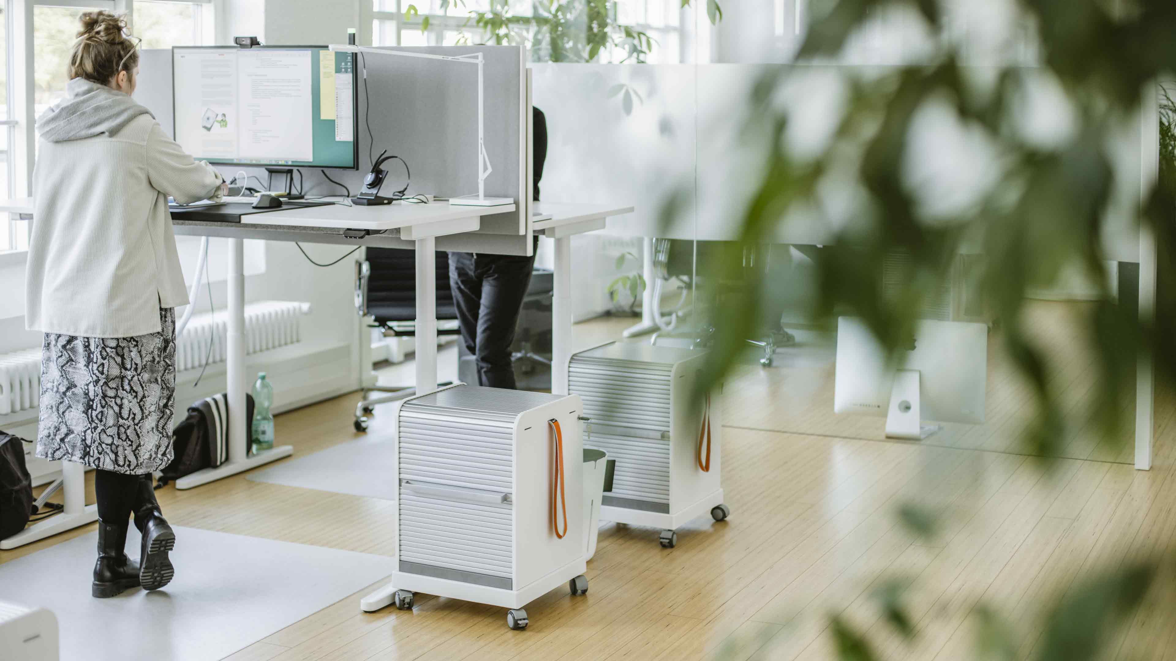 h2_werIst_slider_01.jpg Ansicht eines Arbeitsplatzes am Kieler Standort mit einer Angestellten an einem verstellbaren Stehschreibtisch.