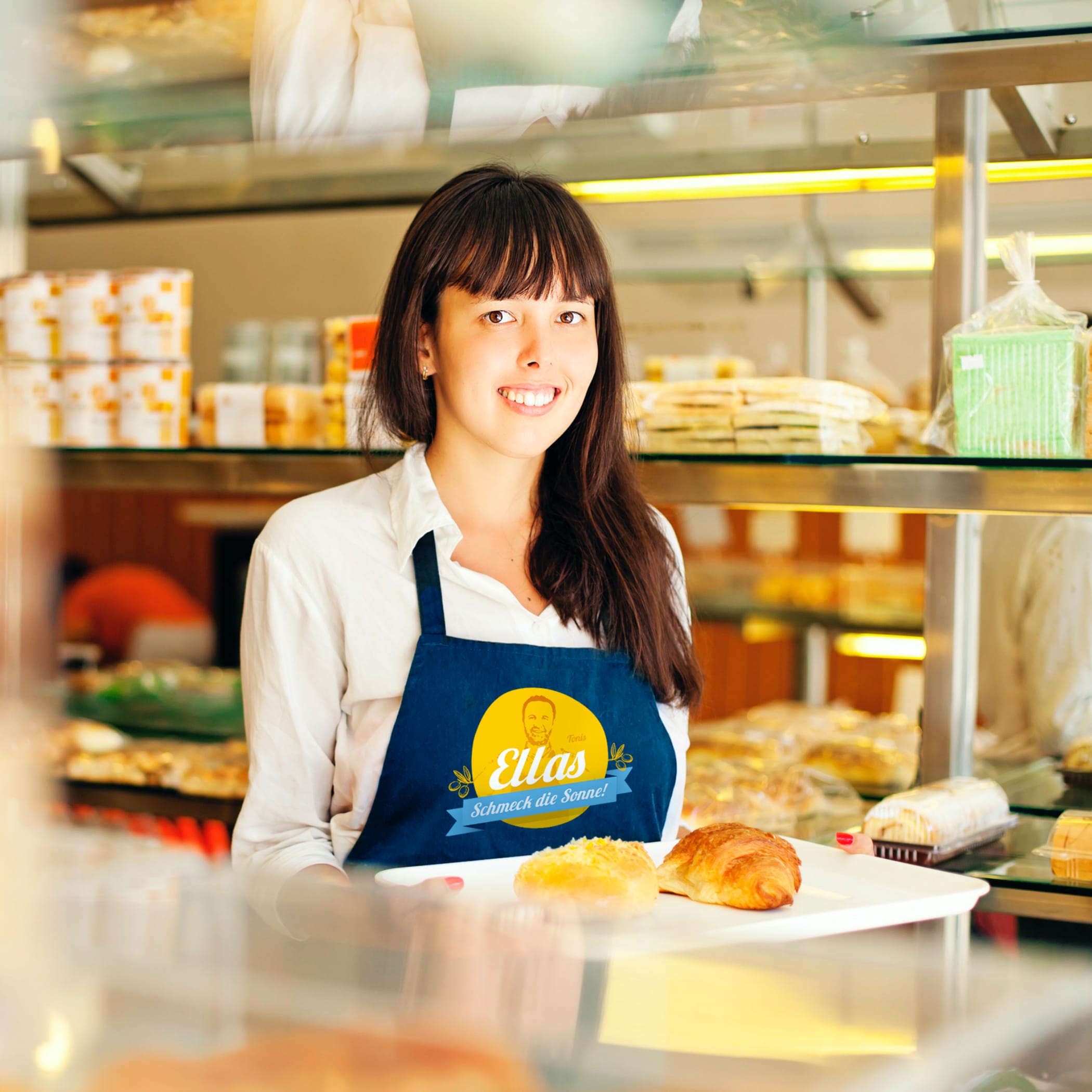 hochzwei_ella_teaser_05.jpg Braunhaarige Mitarbeiterin mit blauer Ellas-Schürze lächelt hinter der Theke