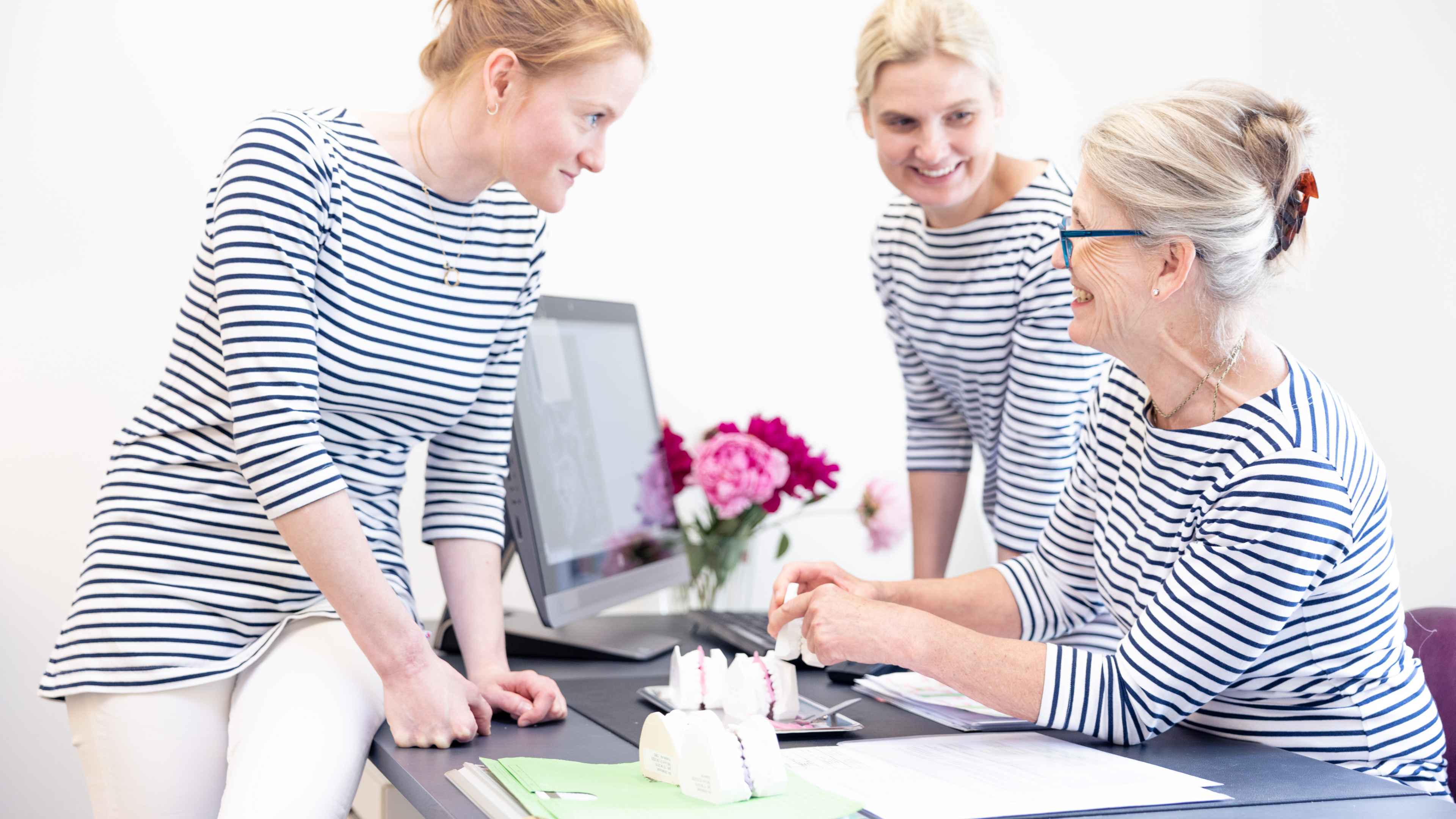 Drei weibliche Angestellte in blau-weiß gestreiften Shirts sprechen an einem Schreibtisch fröhlich miteinander auf dem Schreibtisch liegen Gipsabdrücke von Zähnen