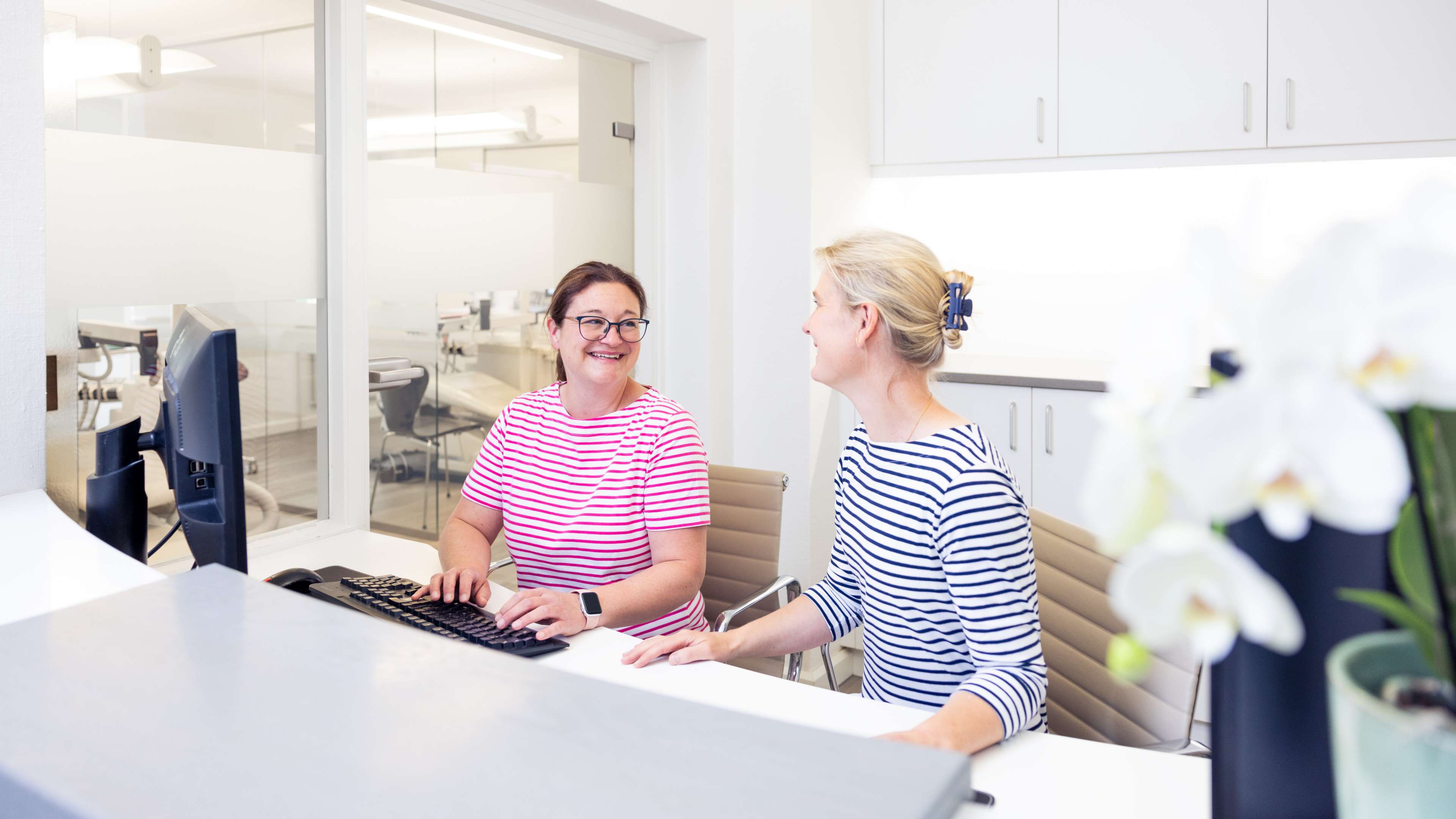 Foto: Zwei weibliche Angestellte sitzen am Empfang der Praxis und sprechen fröhlich miteinander. Die linke Frau trägt ein rot-weiß gestreiftes Shirt, die rechte ein blau-weiß gestreiftes.