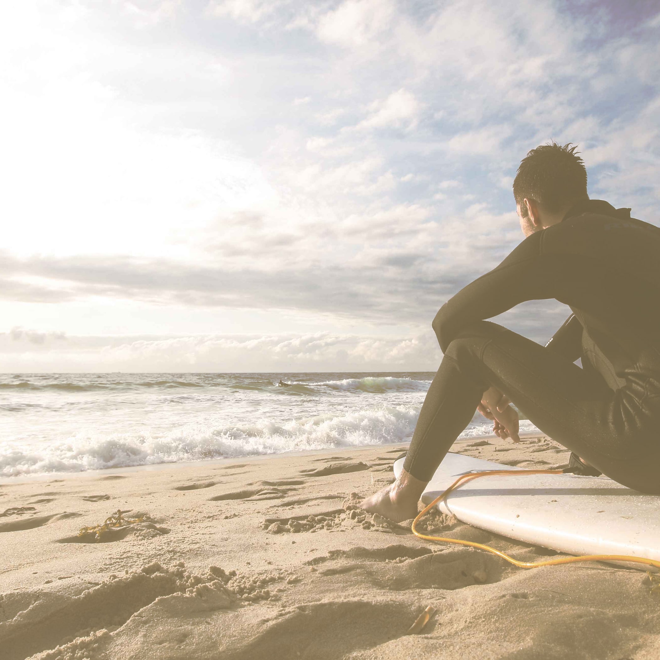 Ein Mann im Neopren Anzug sitzt auf einem Surfbrett am Strand