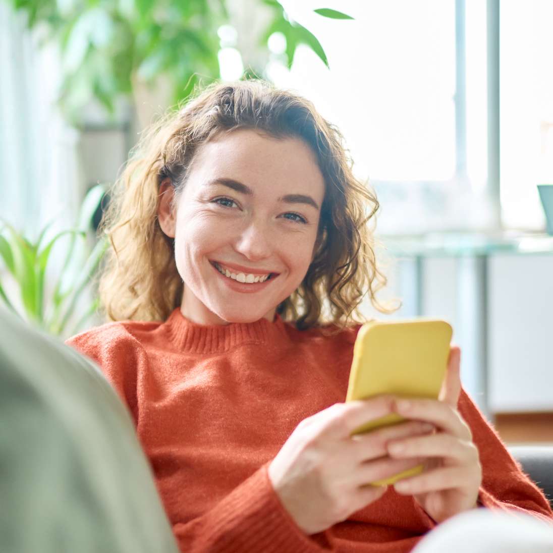 Eine Frau sitzt auf dem Sofa und lächelt in die Kamera. In der Hand ihr Handy.