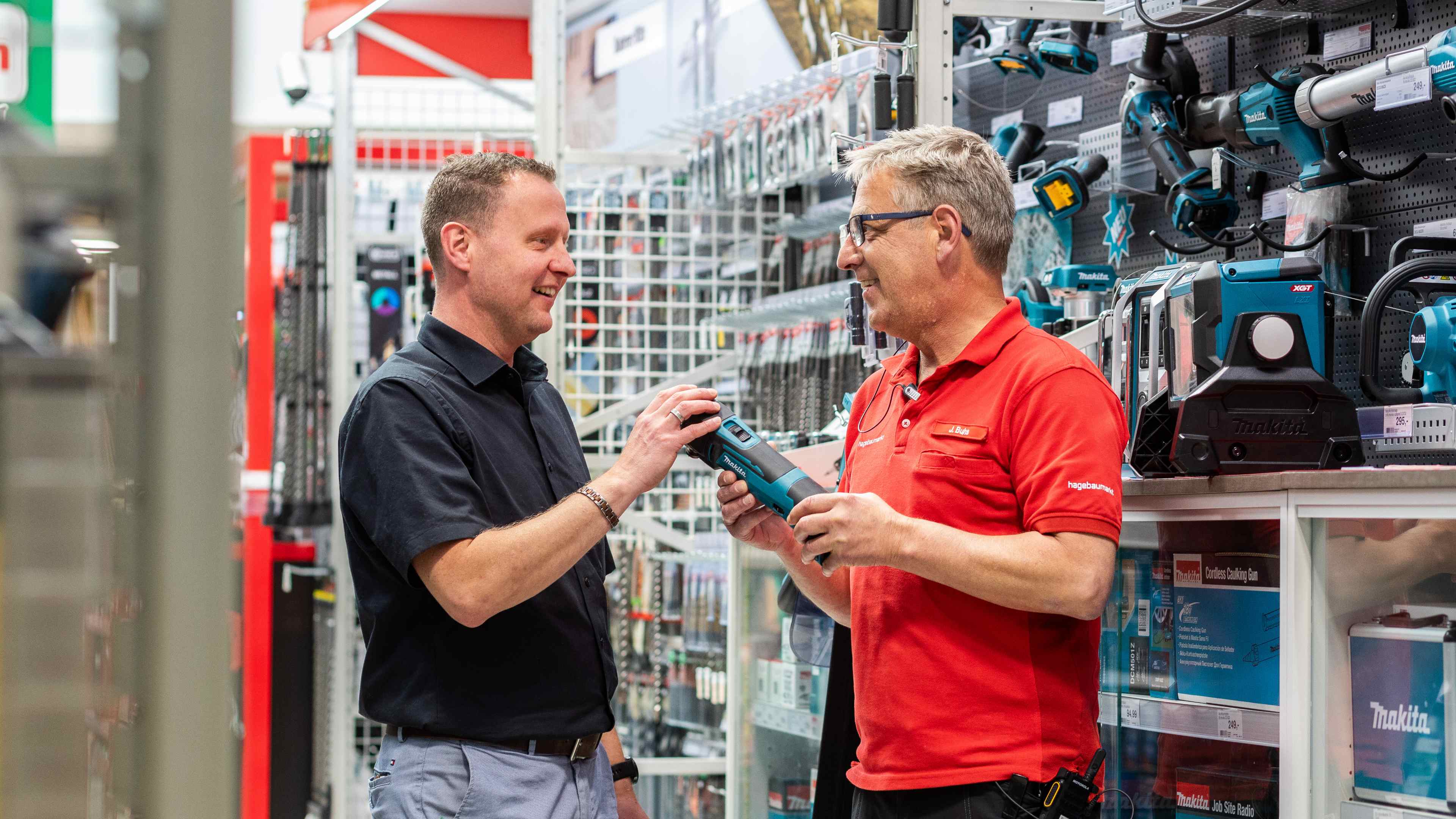 Ein Hagebaumarkt Mitarbeiter in der Makita Abteilung spricht mit einem Kunden und hält dabei ein kleines Gerät von Makita in den Händen.