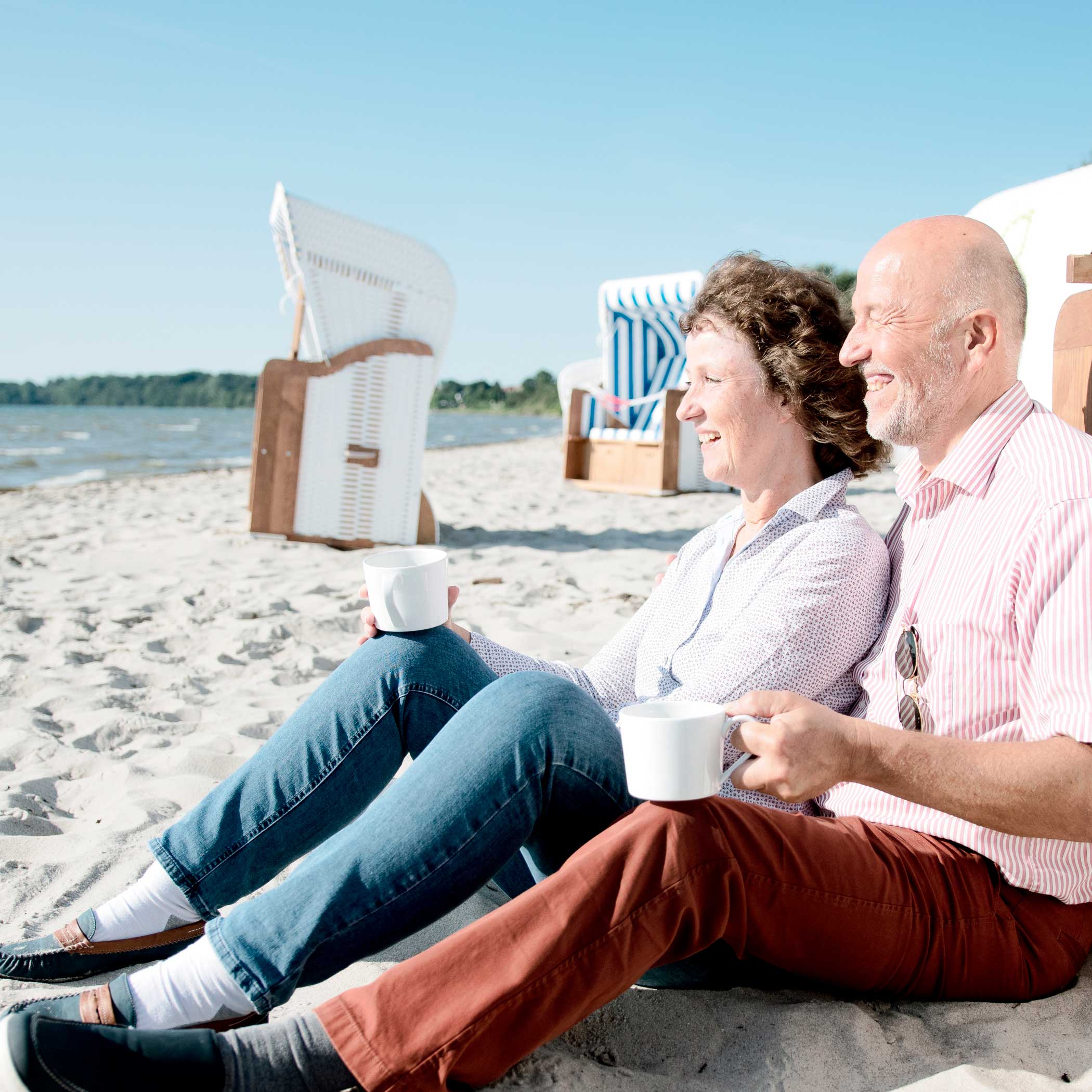 hochzwei_taff2_teaser_03.jpg Älteres Pärchen sitzt mit Tassen am Strand