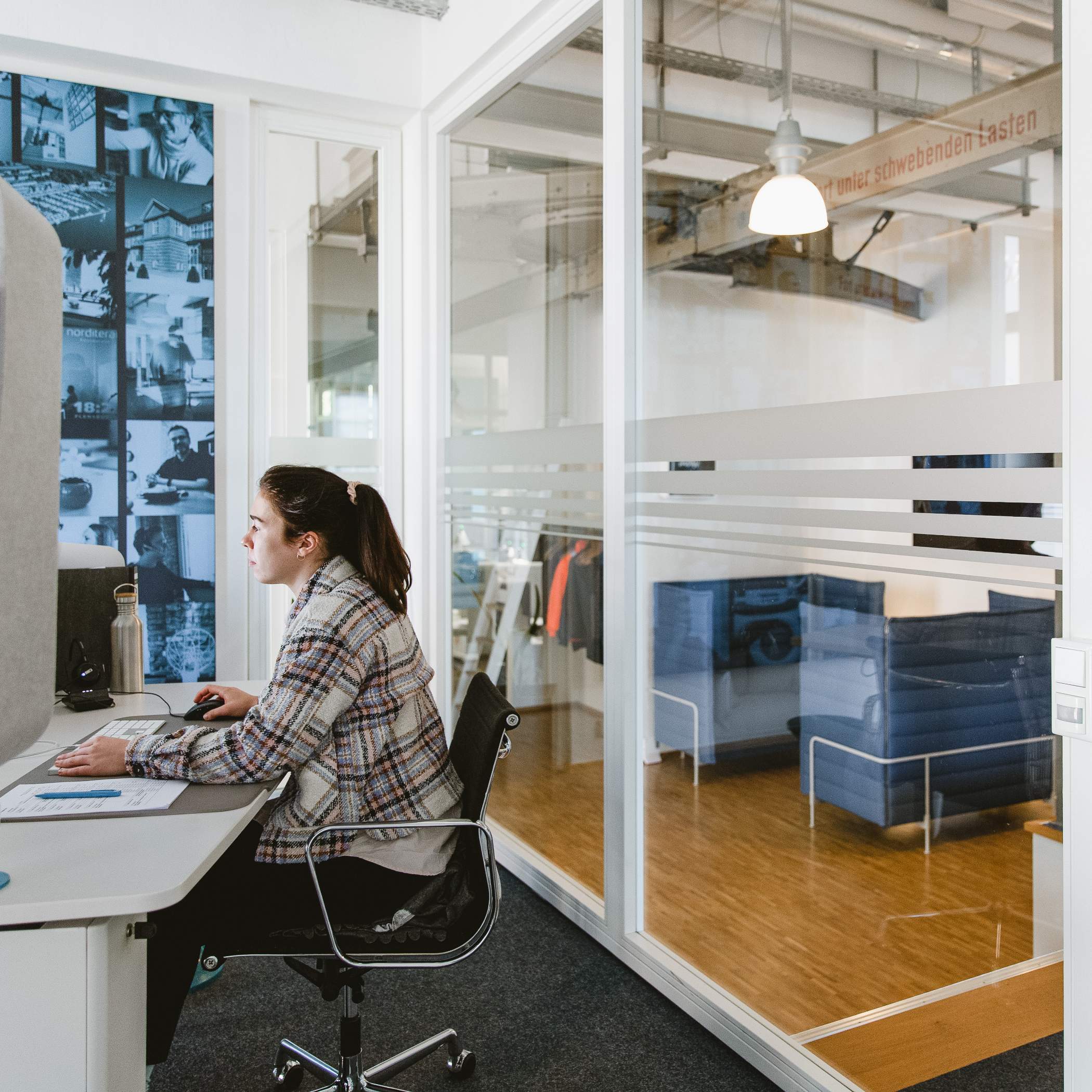 Mitarbeiterin sitzt am Schreibtisch und schaut auf PC, hinter ihr eine Glaswand mit Blick auf eine Sofasitzgruppe im Eingangsbereich