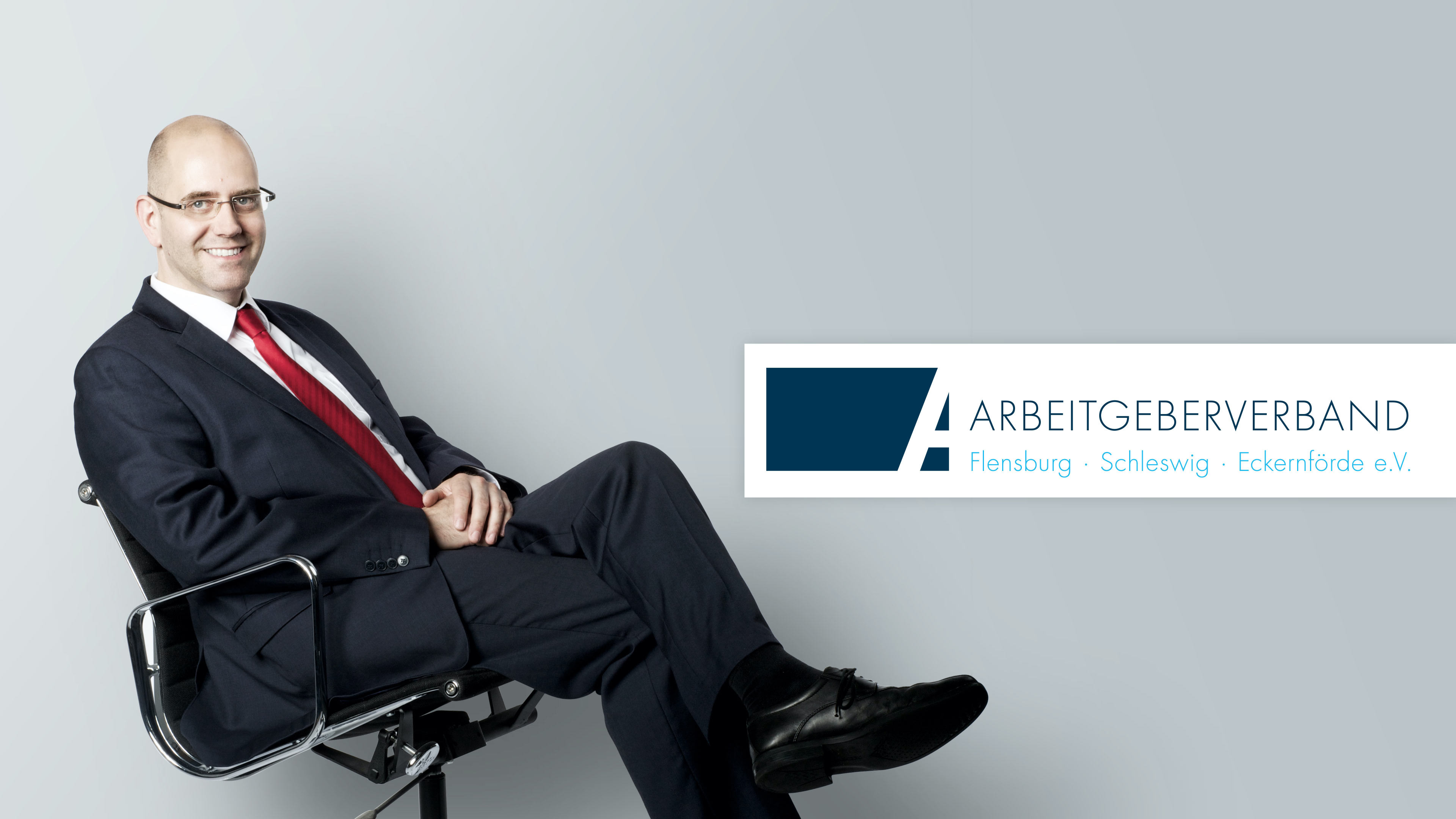 Mann im Bürostuhl und Log des Arbeitgeberverbands vor grauem Hintergrund