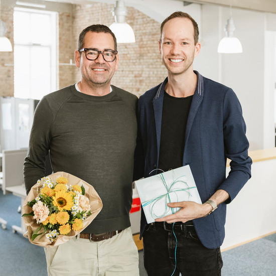 Geschäftsführer Felix und Grafikdesigner Armin posieren mit Blumenstrauß und Geschenk für ein Jubiläumsfoto