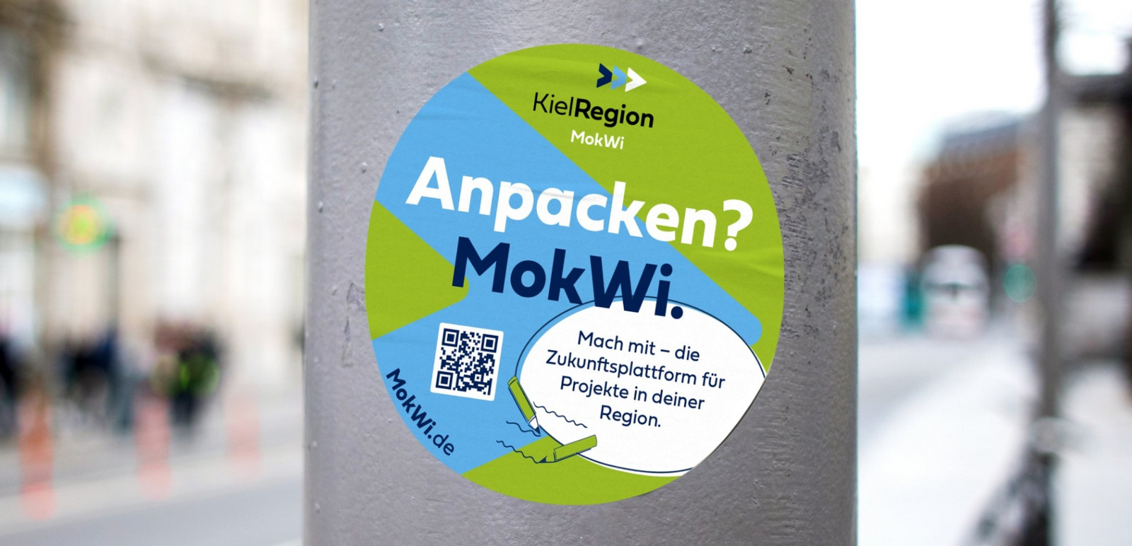 Ei MokWi Sticker auf einem Laternenpfahl. Darauf steht Anpacken? MokWi. Mach mit - die Zukunftsplattform für Projekte in deiner Region. Das abgebildete Logo besteht aus drei Pfeilen nach rechts in dunkelblau, hellblau und weiß.