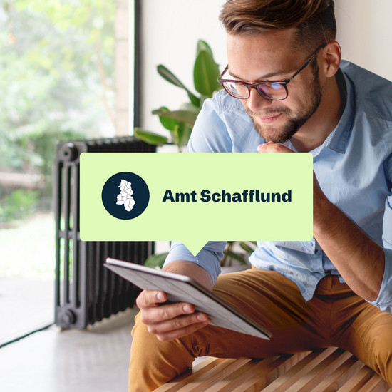 Ein junger Mann sitzt in einem modernen Wohnzimmer und schaut auf ein Tablet. In der Mitte des Bildes ist eine hellgrüne Sprechblase in der Amt Schafflund steht.