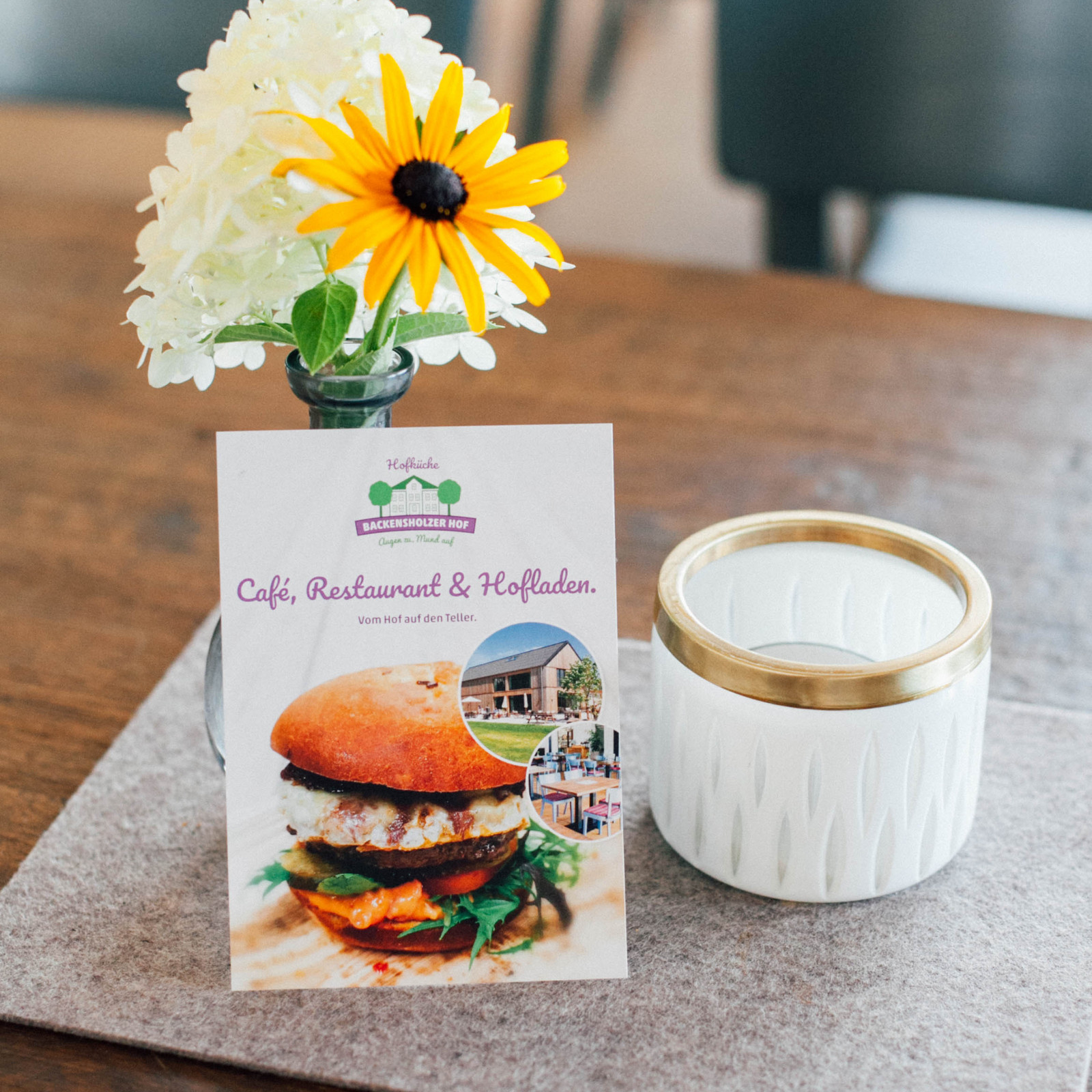 hochzwei_roba_teaser_01.jpg Blumenvase mit zwei Blüten, Teelichthalter und Postkarte mit Backensholzer Logo auf einem Tisch