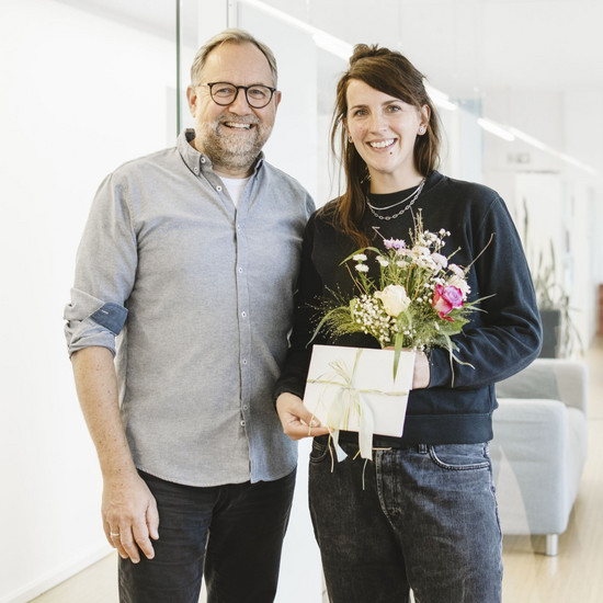 Foto von unserer Kollegin Anni mit Björn, dem Standortschef von Kiel. Anni hält einen Blumenstrauß und ein Geschenk und beide lächeln in die Kamera.