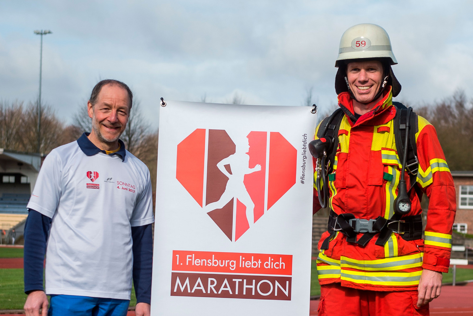 hochzwei_foe_teaser_gross_04.jpg Feuerwehrmann und Mann im weißen T-Shirt neben einem Schild mit Logo und Text