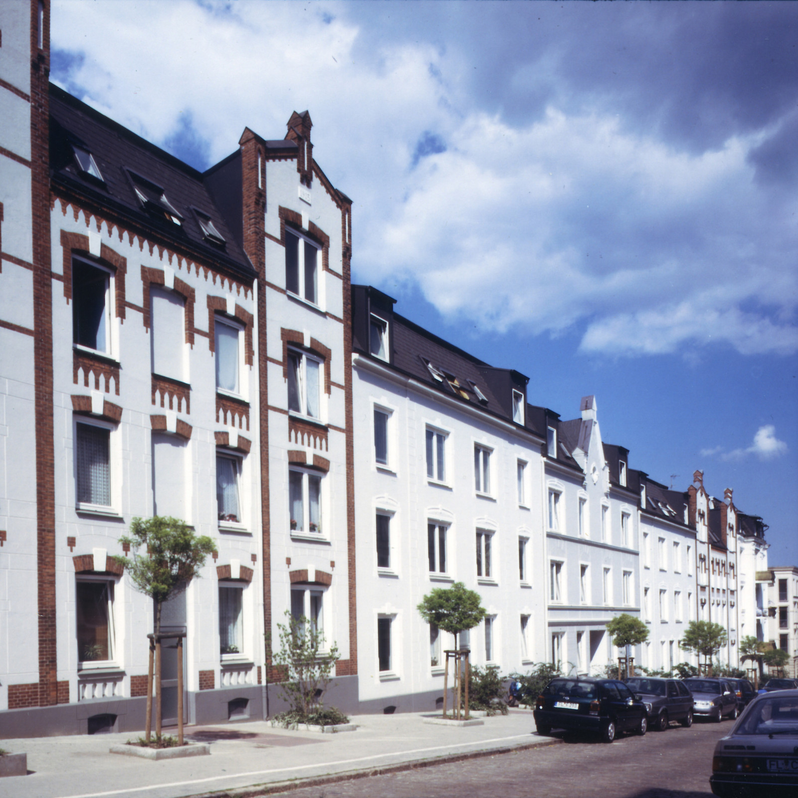 hochzwei_fab_teaser_03.jpg Blick auf dreistöckige weiße Wohnhäuser, davpr kleine Bäume und parkende Autos
