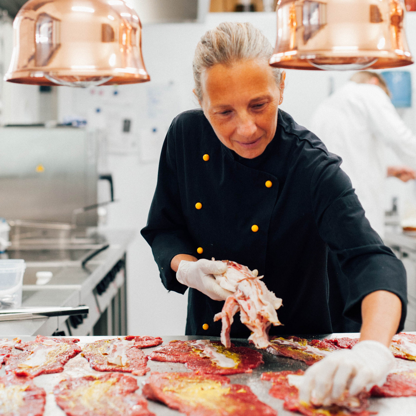 hochzwei_roba_teaser_04.jpg Eine schwarz gekleidete Köchin verteilt Speck auf vor ihr ausgebreiteten Rouladen auf einem Tisch