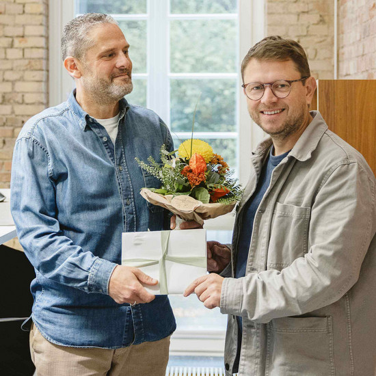 Foto von Chef Timo und Grafiker Leo. Timo überreicht Leo ein Geschenk und einen Blumenstrauß.