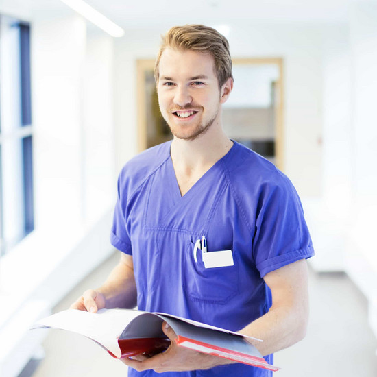 Mann im lila Pflegehemd mit Mappe in der Hand lächelt in die Kamera