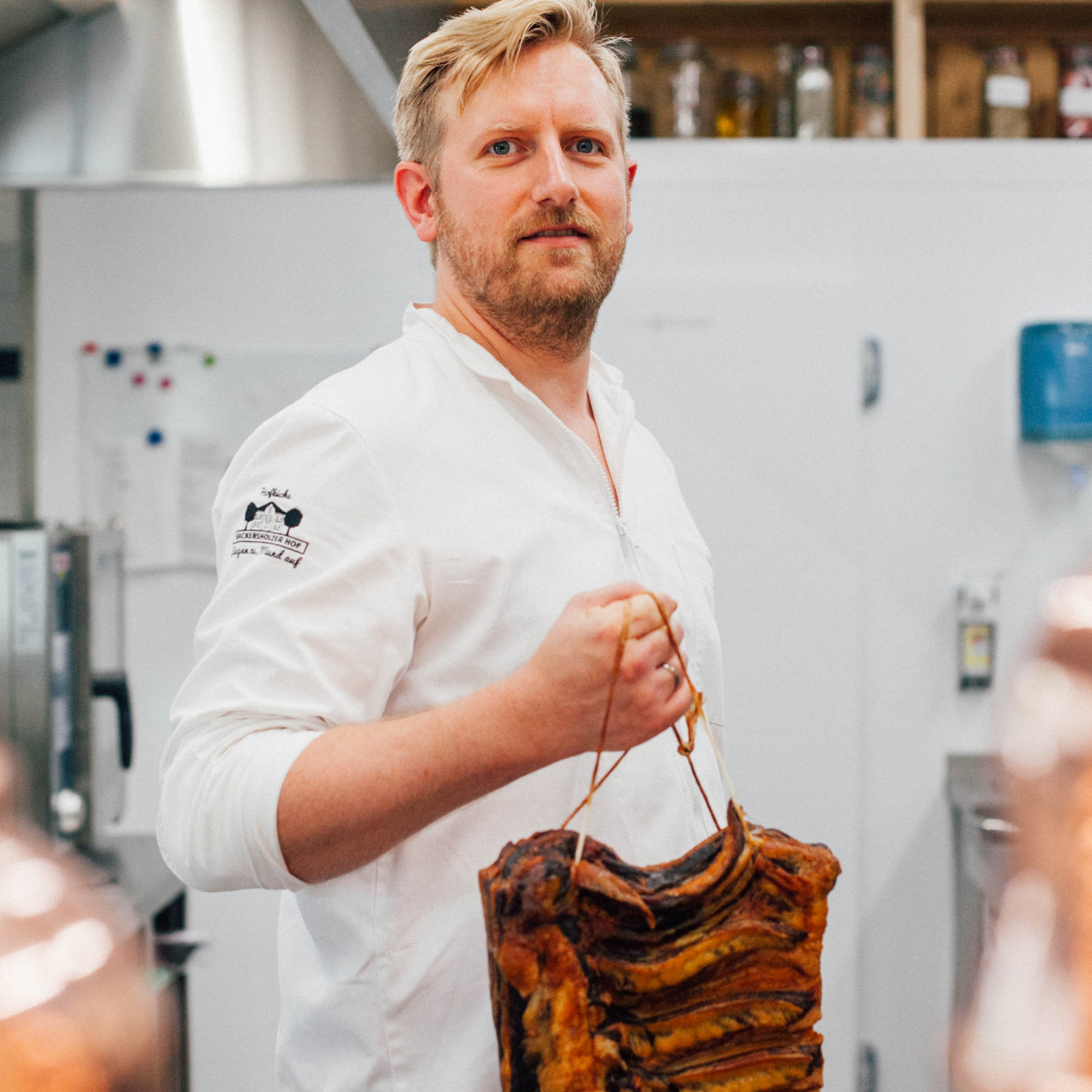 hochzwei_roba_teaser_12.jpg Bärtiger Mann im weißen Oberteil hält ein großes Stück Fleisch mit Rippen