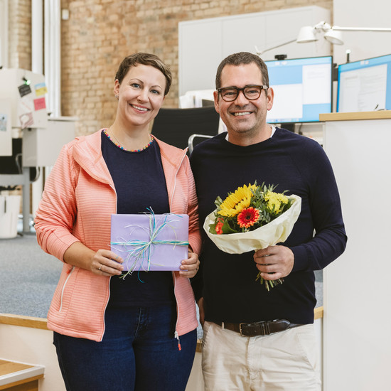 Foto unserer Kollegin Rebecca mit Chef Felix. Rebecca hält ein verpacktes Geschenk und Felix hält einen Blumenstrauß.