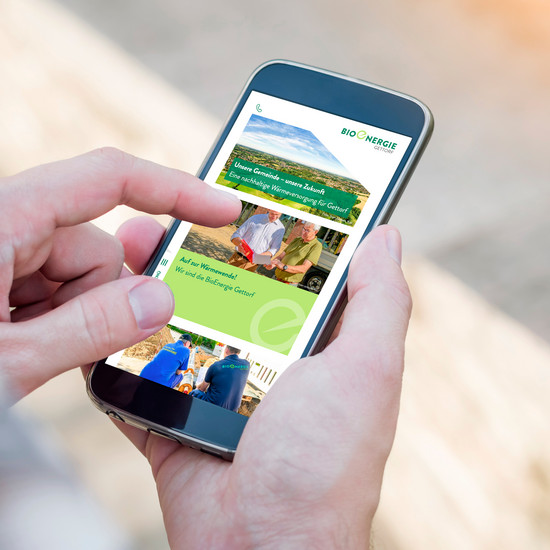 Wärme ist grün … Zwei Hände halten Smartphone, das Website von Bioenergie Gettorf zeigt