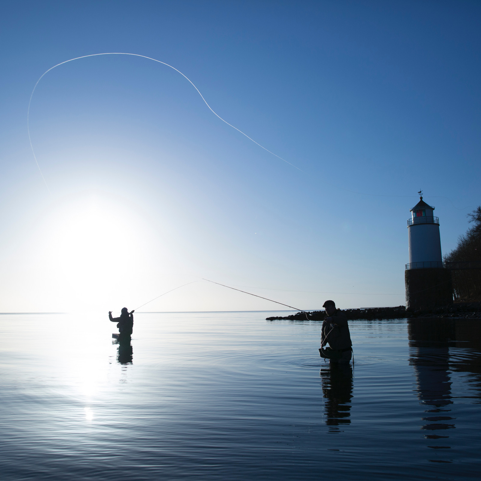 hochzwei_deso_teaser_08.jpg Zwei Fliegenfischer in Wathosen stehen im Wasser vor blauem Himmel, kleiner Leuchtturm rechts