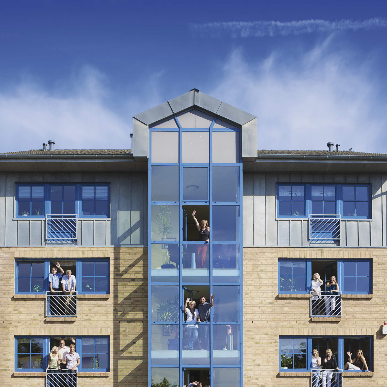 hochzwei_fab_teaser_02.jpg Blick auf gelbes Wohnhaus mit blauen Fenstern, aus welchen fröhliche Bewohner winken