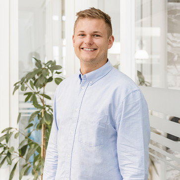 Porträtfoto von Tom er trägt ein hellblaues Hemd und lächelt freundlich in die Kamera. Im Hintergrund Glaswände der Büroräume und eine Pflanze.