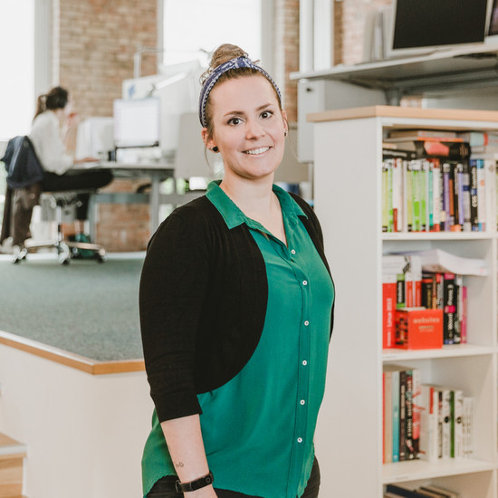 Portraitbild von Henrike in den Räumlichkeiten von Hochzwei in Flensburg. Sie lächelt freundlich in die Kamera und trägt eine grüne Bluse mit schwarzem Bolerojäckchen darüber. Ihre Haare sind hochgesteckt.