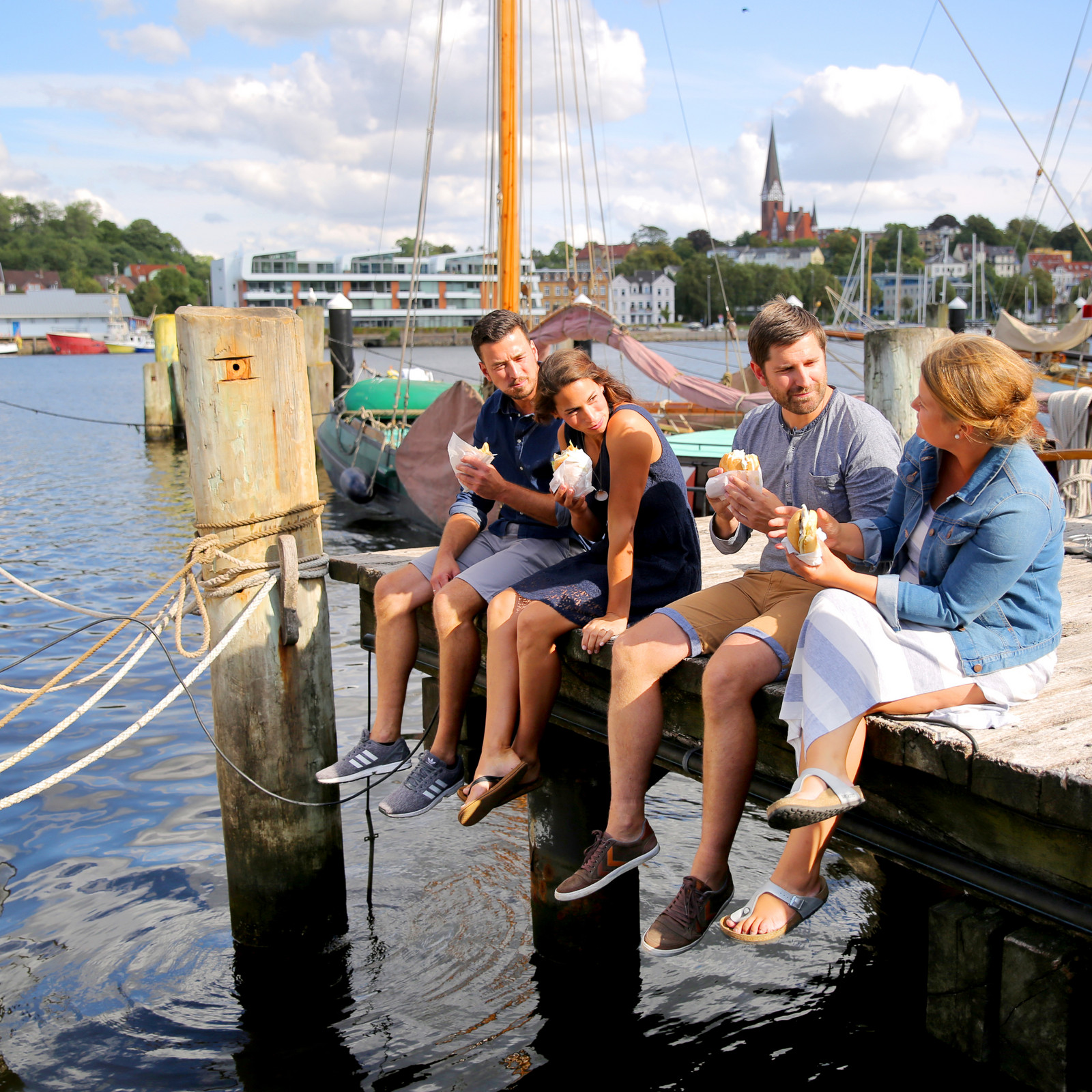 hochzwei_taff_teaser_03.jpg Vier Personen sitzen auf einem Steg und essen ein Fischbrötchen