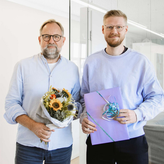 Standortchef Björn mit Kollege Martin. Sie lächeln freundlich in die Kamera und halten einen Blumenstrauß und ein verpacktes Geschenk.
