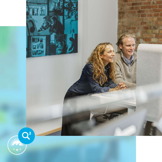 Festanstellung Zwei HOCHZWEI-Mitarbeiter stehen vor einem Arbeitsplatz und blicken auf Bildschirm