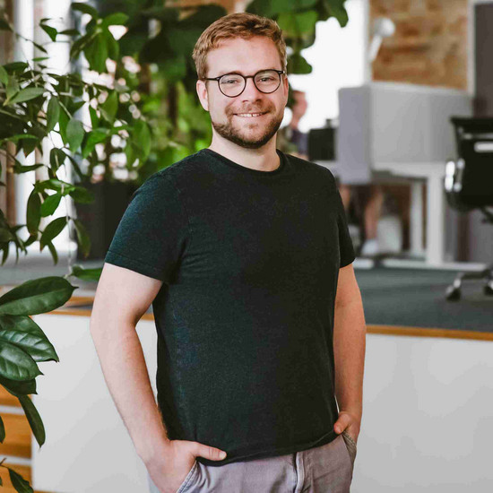 Porträit von Bryan. Er trägt eine Brille und ein dunkles T-Shirt und lächelt in die Kamera.